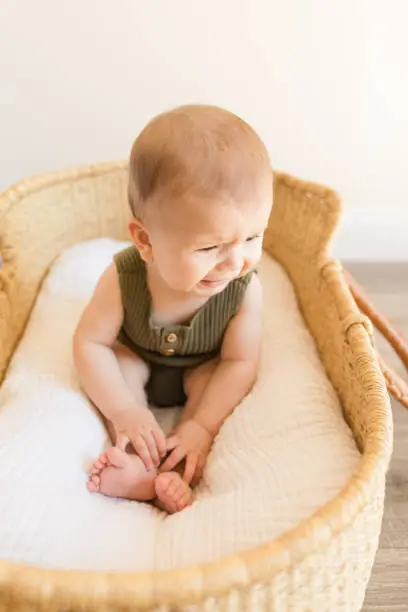 A Cranky 27-Week-Old Baby Boy Wearing an Olive Green Knit Romper While Sitting in a Cozy Cotton Blanket in a Seagrass Moses Basket in a Neutral Modern Home and Playing.