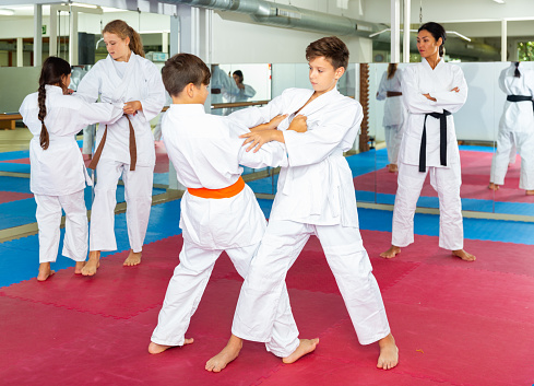 Kids training in gym during group karate classes. Female trainer standing beside and watching them.