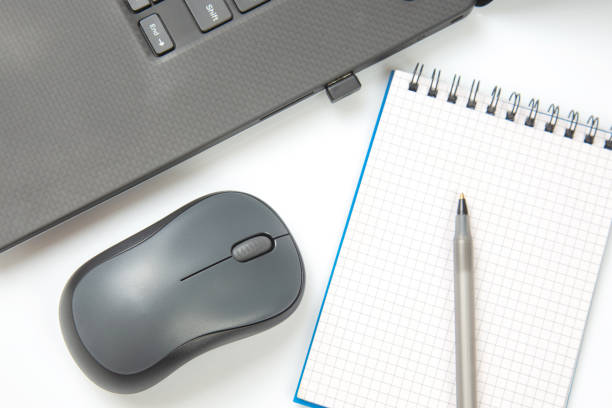 istock Computer wireless mouse, pen and notepad next to a laptop on a white table close-up. Items for office work 1407386099