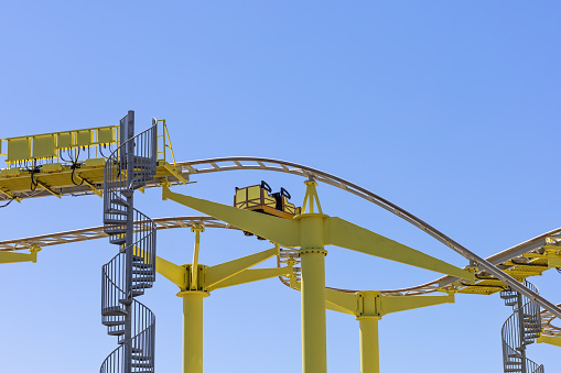 A yellow slide with an empty booth and spiral staircase leading to it against a blue sky. A non-working attraction in an amusement park. Service ladder.