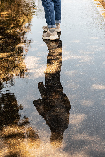 Legs of woman in jeans and sneakers. The silhouette of a woman is reflected in a puddle on the pavement. Walk after the rain.