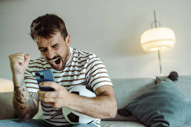 Sports betting and the joy of winning Excited young man watching sports game and cheering for his team, He is holding mobile phone and he is happy about winning the bet gambling stock pictures, royalty-free photos & images