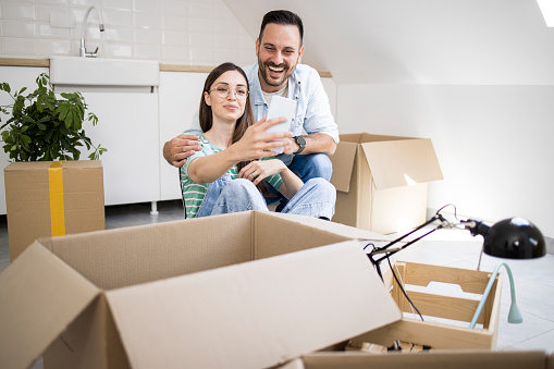 Happy young couple moving into a new home, unpacking their belongings and using smartphone.