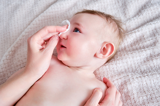 Las manos de la madre lavan la cara de un bebé feliz con una almohadilla de algodón. Mamá sostiene higiene niño pequeño sonriente en el sofá, de seis a siete meses de edad photo