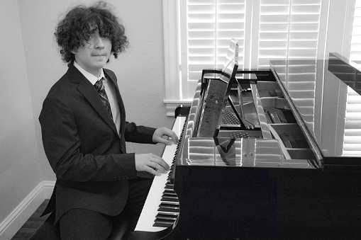 A teen boy plays and composes on a baby grand piano. Black and white.