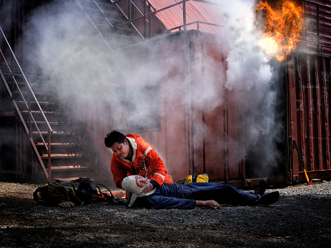 1 Fireman weraing heat protection suit perform cardiopulmonary CPR heart pump to friend lay unconciousness from the smoke and fire burn in container background