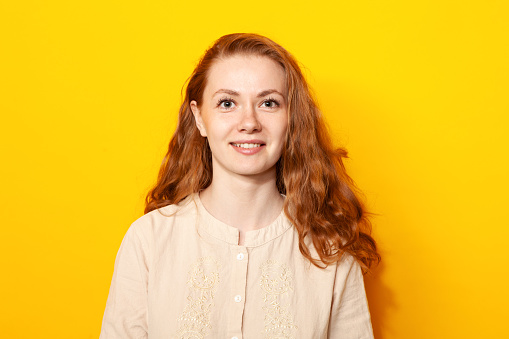 Close-up studio portrait of an attractive 24 year old red-haired woman in a beige blouse on a yellow background