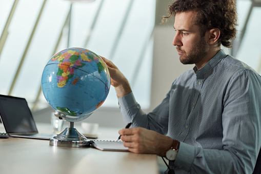 Young businessman examining the globe while taking notes in the office.