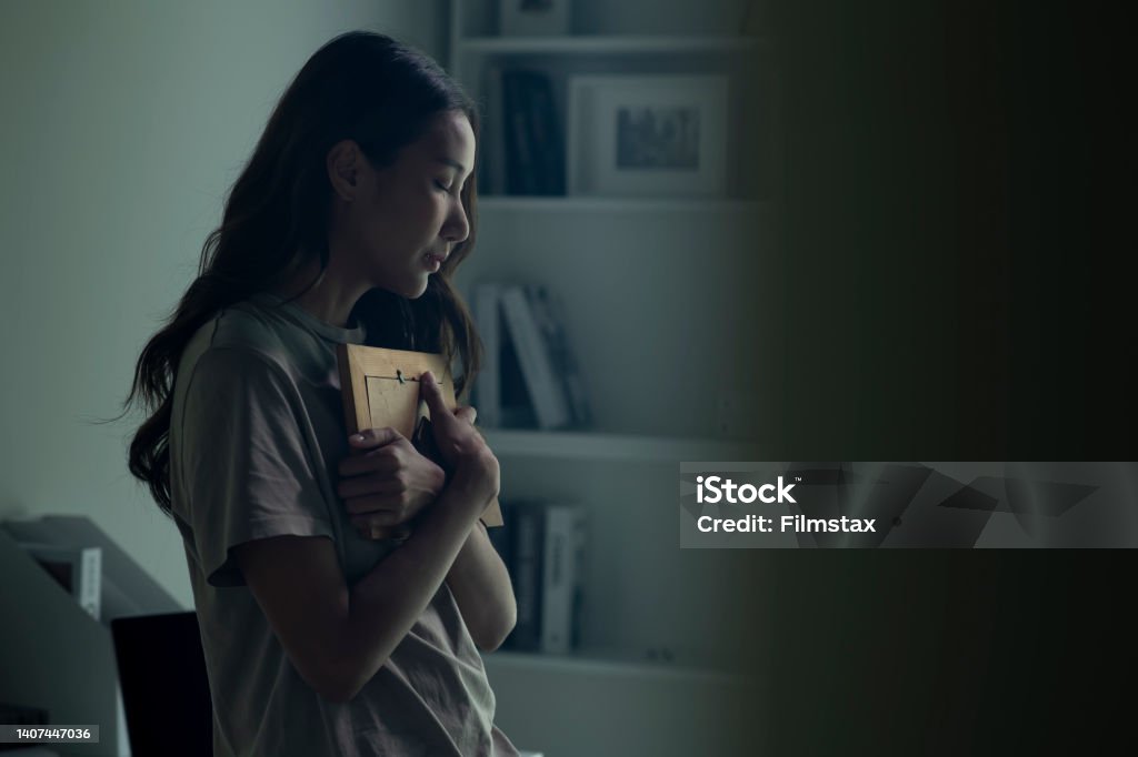 Asian women holding a photo frame of lost loved one and crying. Woman feeling sad when looking at picture of lost loved one in the frame. Depressed Asian women holding a photo frame of lost loved one and crying. Grief Stock Photo