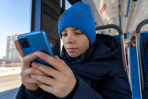 Boy sits at window and looks with interest at screen of smartphone. Happy caucasian kid using cell playing mobile games online on smartphone connected to public wifi sitting on seat in city tram.