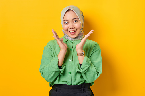 Cheerful young Asian woman in green shirt celebrating success on yellow background