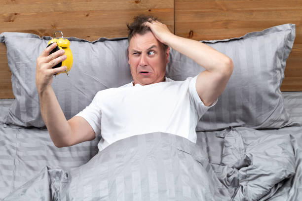 l'uomo scontento giace a letto con una maglietta bianca e guarda una sveglia gialla che tiene in mano. concetto quando ho dormito troppo per lavorare - dormire troppo foto e immagini stock