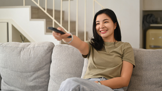 Happy young woman holding remote control, watching tv on couch, enjoying carefree leisure weekend activity at home.