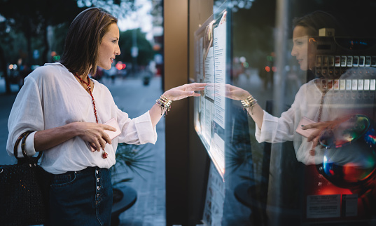 Side view of concentrated mature lady in trendy outfit standing near window of cafe and reading menu while walking on city in evening