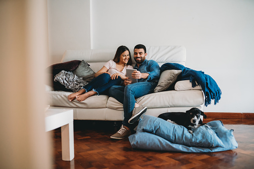 Young adult couple together at home. They are sitting on the sofa using a tablet together, watching a tv show. Their dog is lying down near them.