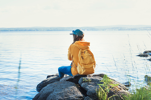 rear view Young woman wearing cap, yellow hoodie with backpack sitting on rocks and looking at view of lake or river Enjoying nature