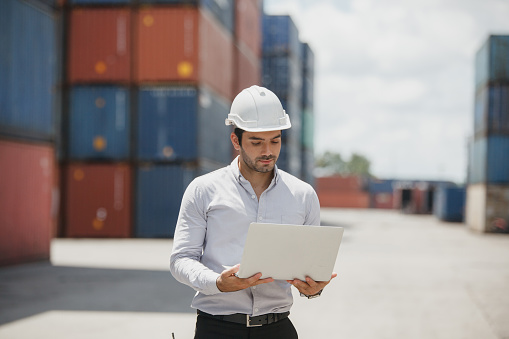 Engineer worker in causual suit standing in shipping container yard holding laptop with smile. Import and export product. Manufacturing transportation and global business concept