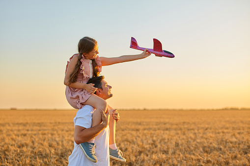 Cute girl riding on father's shoulder and playing with toy airplane against sky in wheat field