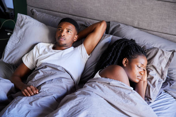 jovem parecendo infeliz enquanto estava deitado na cama com sua esposa dormindo - tired men african descent sadness - fotografias e filmes do acervo