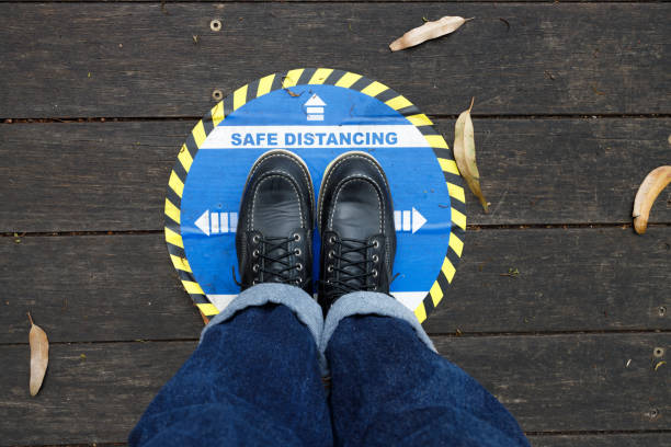 A woman's feet standing at a designated safe social distancing mark taped onto a wooden floor.