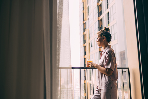 A side view of a pensive Asian female in pajamas drinking orange juice while standing on the balcony of her hotel room early in the morning.