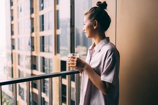 A side view of a happy Asian female in pajamas drinking orange juice while standing on the balcony of her hotel room early in the morning.