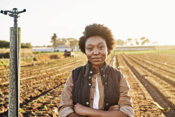 晴れた日に農場でコートを着た若いアフリカの女性農家。ヒスパニック系労働者は、カジュアルな服装で手を組んで畑に立っています。農場でポーズをとってカメラを見つめる黒人女性。 - africa farmer african descent agriculture ストックフォトと画像