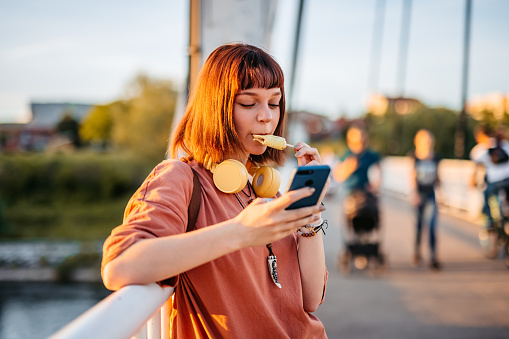 Beautiful young woman using smart phone and eating ice cream on the bridge on a sunny day.