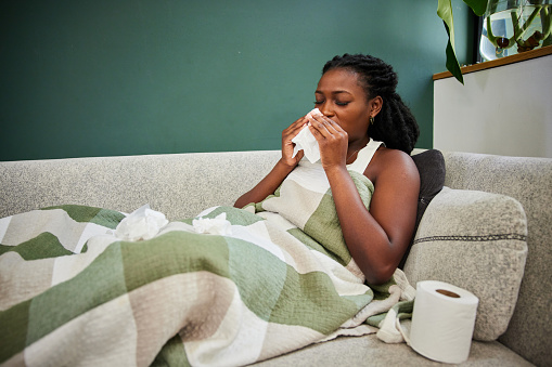 Young African woman with a cold sneezing into tissue while lying wrapped in a blanket on her living room sofa