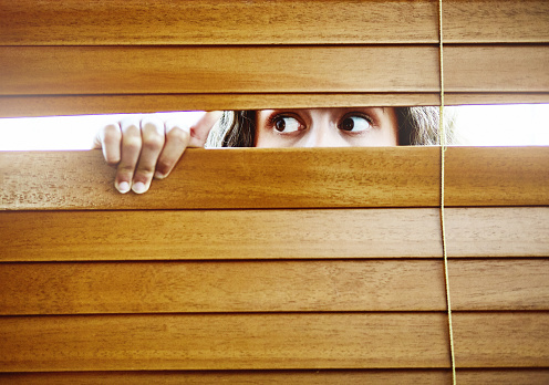 Snooping or fearful, a young woman looks out through a window, parting the blinds with her hand.