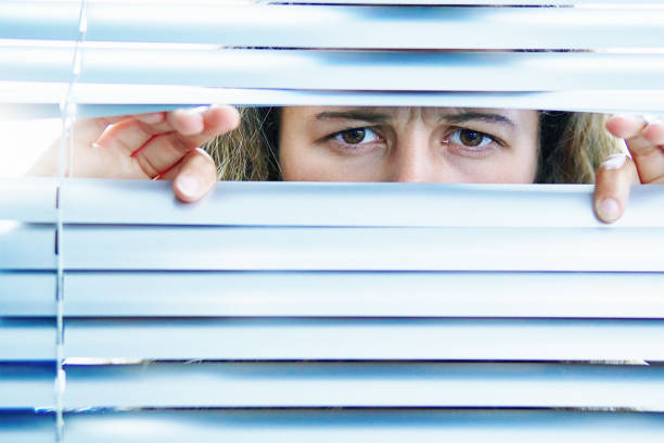 istock Anxious young woman peers furtively through window blinds 1407502406
