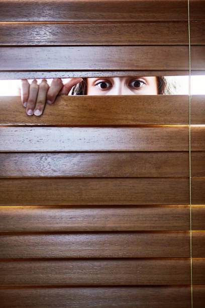 istock Anxious young woman peers furtively through window blinds 1407502468
