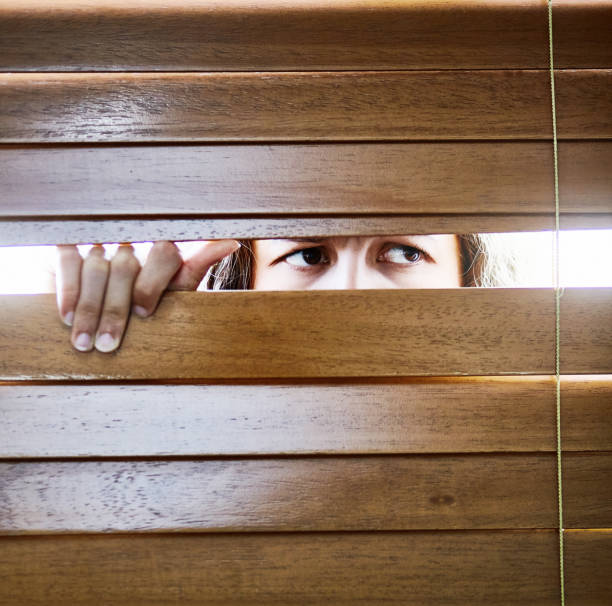 istock Anxious young woman peers furtively through window blinds 1407502517
