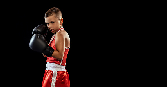 Young beginner boxer, sportive boy training isolated over dark background. Concept of sport, movement, studying, achievements lifestyle. Kid practicing punch. Copy space for ad
