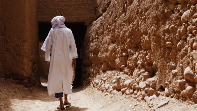 A traditional dressed Moroccan man wearing a white gandoura and a turban walks inside a Kasbah in Tamnougalt, Morocco.