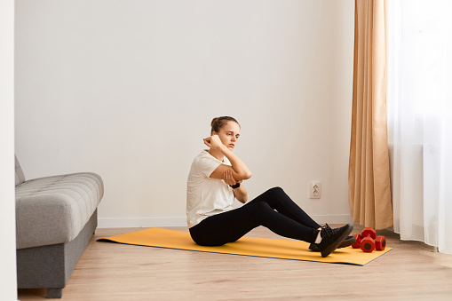 Side view of sick woman sitting on yoga mat feeling elbow pain while doing exercise for good body shape at home, touching painful place, frowning face, being injured during workout.