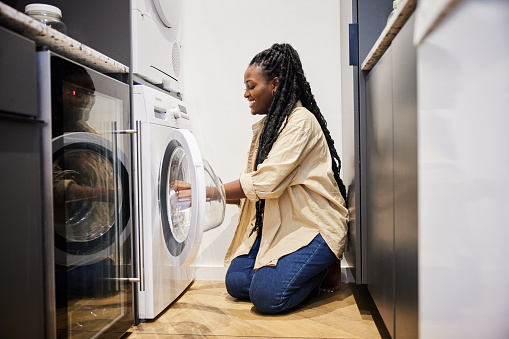 Smiling young African woman kneeling on the floor of a utility room and putting clothing into a washing machine while doing housework