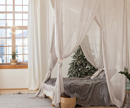 Christmas scandinavian bedroom with white canopy with lights. Christmas tree and bed in cozy light room