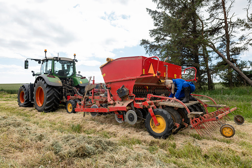 A female farmer wearing overalls and wellington boots crouching down on the back of a stationary seed drill in a field on a sustainable farm in Embleton, Northumberland. She is examining the machine and checking everything is in working order. She is preparing to use the drill to drill and sew the field with pollinators to produce cover crops to encourage wildlife to the farm.