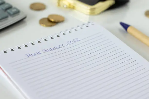 Handwritten text "home budget 2022" on a blank notebook with pen, calculator, golden coins, and a money purse on white. Family budgeting, finance plan, expense, and saving calculation concept.