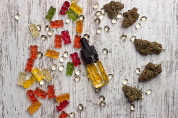 A glass bottle with yellow cannabis cbd oil lies on a wooden table surrounded by dry medical marijuana buds, cbd extract capsules and colorful cbd gummy bears. A glass bottle with yellow cannabis cbd oil lies on a wooden table surrounded by dry medical marijuana buds, cbd extract capsules and colorful cbd gummy bears. On a wooden background, top view bud candy stock pictures, royalty-free photos & images