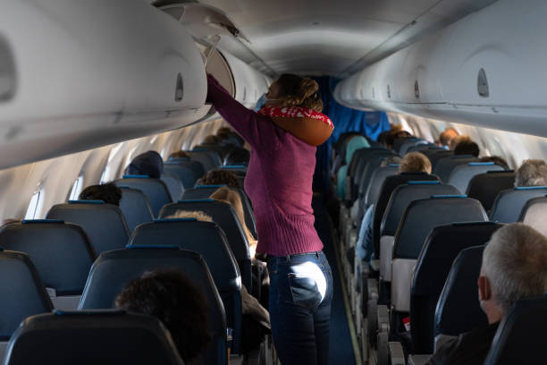 Woman placing suitcase inside overhead compartment in plane Woman with neck pillow standing on airplane aisle placing suitcase inside overhead compartment. Passenger taking carry on luggage on plane economy class stock pictures, royalty-free photos & images