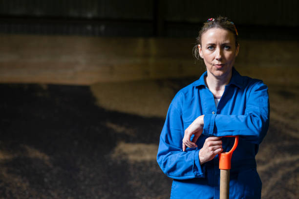 Female Blue Collar Worker Portrait A close up of a female blue collar worker stood looking into the camera leaning on a shovel as she stands proudly in a barn full of rapeseed and wheat. outhouse interior stock pictures, royalty-free photos & images