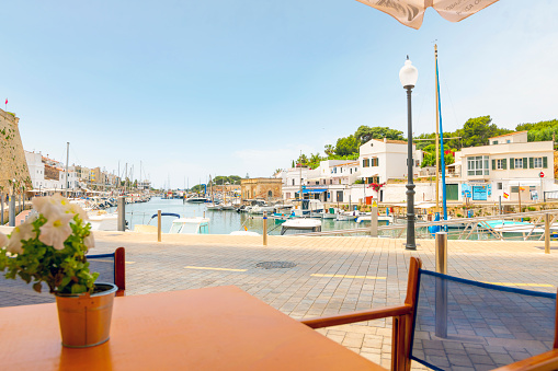 View from a waterfront sidewalk cafe table of the colorful harbor filled with boats in the historic city of Ciutadella de Menorca, Spain, on the island of Menorca, Spain.