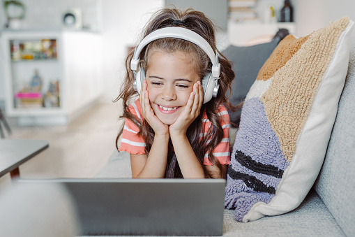 A cute little girl uses a laptop at home in the living room. She is lying down on the sofa and has wireless headphones on her head while watching online content.