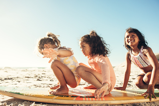 Exploring with a surfing board at the beach. Three happy little girls playing on a surfing board on sea sand. Group of adorable little children having fun together during summer vacation.