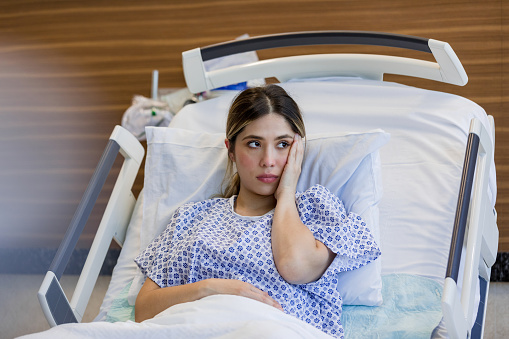 Lying on the hospital bed, the emotionally distressed young adult woman looks out the window with her hand on her face.