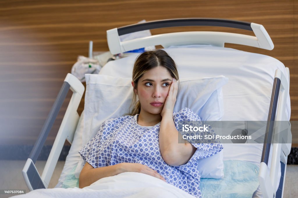 Sad young adult woman in hospital puts hand to face Lying on the hospital bed, the emotionally distressed young adult woman looks out the window with her hand on her face. Emergency Room Stock Photo