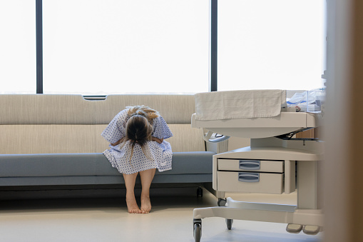 The young adult woman bends forward, puts her head in her lap and weeps in the hospital room.  In the foreground is an infant warmer.