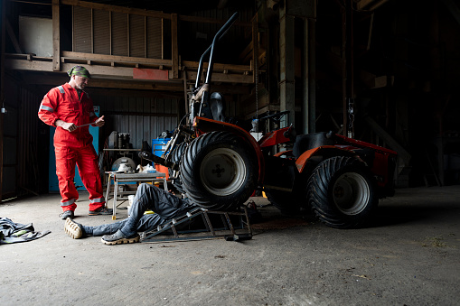 A wide angle view of two farm workers who are working on a tractor together in a barn where they spotting each other and helping by handing tools to each other.